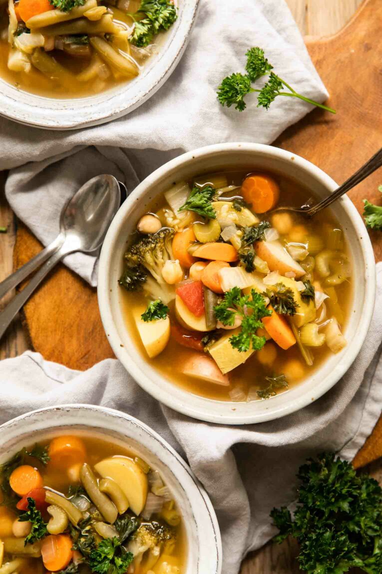 Top shot of 3 bowls of vegetable soup with spoons and fresh parsley.